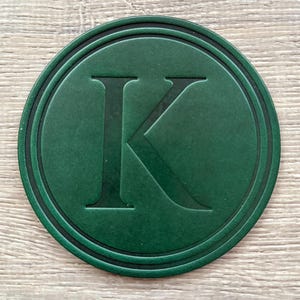 Könnte beinhalten: Ein runder, dunkelgrüner Untersetzer mit einem geprägten Buchstaben "K" in der Mitte. Der Untersetzer hat eine strukturierte Oberfläche und einen schwarzen Rand, vor einem hellen Holzmaserungshintergrund. Ca. 10 cm Durchmesser.
