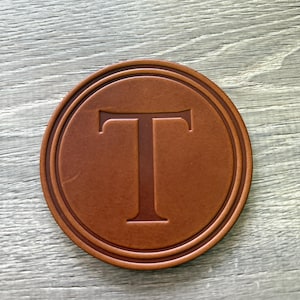 Puede incluir: Un posavasos redondo de cuero marr&oacute;n con la letra "T" grabada en el centro. El posavasos tiene un borde en relieve y est&aacute; sobre una superficie de madera clara. Un dise&ntilde;o simple y elegante.