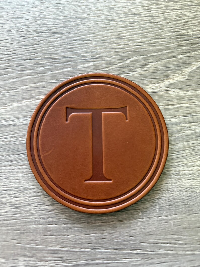Puede incluir: Un posavasos redondo de cuero marr&oacute;n con la letra "T" grabada en el centro. El posavasos tiene un borde en relieve y est&aacute; sobre una superficie de madera clara. Un dise&ntilde;o simple y elegante.