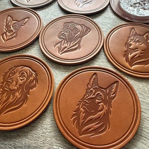 May include: A collection of brown leather coasters, each featuring a detailed embossed image of a different dog breed. The coasters are round with a raised edge, and the dog portraits include a pug, German Shepherd, and others.