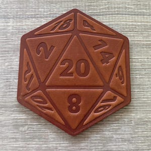Puede incluir: Un dado D20 de cuero marrón con números en relieve. El dado de veinte caras presenta los números 2, 4, 8, 9, 12, 14, 16, 17, 18 y 20. Es un accesorio de juego.