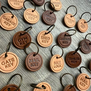 Peut inclure: Plusieurs porte-clés ronds en cuir, de couleurs marron et beige, avec des inscriptions en relief telles que "Garage Key" et "Office Key". Chaque porte-clé est équipé d'un anneau métallique.