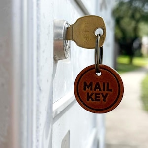 May include: A brass key inserted into a mailbox lock. The key has the number "3748PS" stamped on it. A brown leather keychain tag with the words "MAIL KEY" is attached to the key.