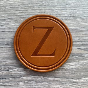 May include: A round, brown leather coaster with a debossed letter "Z" in the center. The coaster has a double-lined border and is set against a gray wooden surface. The coaster is a personalized item.