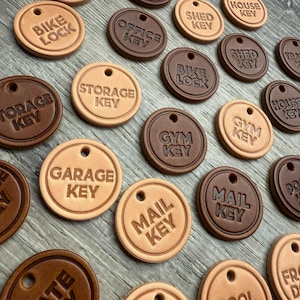 May include: A collection of round, leather keychains in shades of brown and tan. Each keychain is embossed with a different key label, such as "Bike Lock," "Garage Key," and "Mail Key." The keychains have a small hole for attachment.