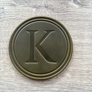 Puede incluir: Un posavasos redondo de color bronce con un borde en relieve y una gran letra "K" en el centro. El posavasos tiene un aspecto ligeramente envejecido, lo que sugiere un estilo vintage. El fondo es una veta de madera clara.