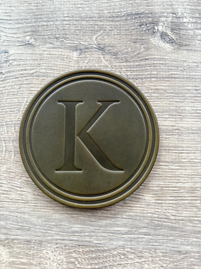 Puede incluir: Un posavasos redondo de color bronce con un borde en relieve y una gran letra "K" en el centro. El posavasos tiene un aspecto ligeramente envejecido, lo que sugiere un estilo vintage. El fondo es una veta de madera clara.