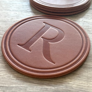 Puede incluir: Un conjunto de posavasos de cuero marrón con la letra "R" en relieve en el centro. Los posavasos tienen forma circular con un borde elevado. El color es marrón rojizo cálido.
