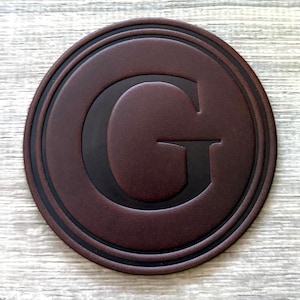 May include: A round, brown leather coaster with a raised black letter "G" in the center. The coaster has a dark brown border with two concentric rings. The overall design is simple and elegant.