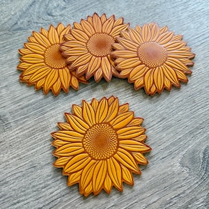 May include: A collection of sunflower-shaped coasters. The coasters are a warm yellow-orange color with detailed petal and center designs. The coasters are made of a durable material and are ideal for protecting surfaces from drink rings.