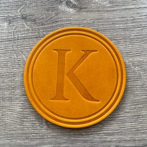 Puede incluir: Un posavasos redondo naranja con un borde en relieve y una letra "K" may&uacute;scula en el centro. El posavasos est&aacute; hecho de un material s&oacute;lido y tiene una superficie texturizada. El dise&ntilde;o es simple y elegante.
