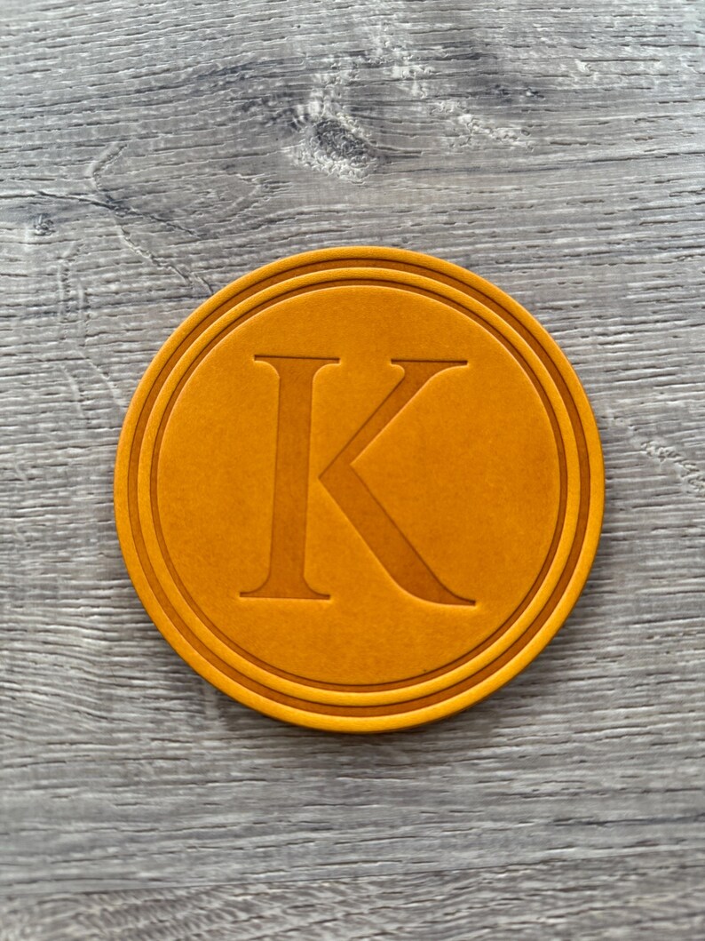 Puede incluir: Un posavasos redondo naranja con un borde en relieve y una letra "K" may&uacute;scula en el centro. El posavasos est&aacute; hecho de un material s&oacute;lido y tiene una superficie texturizada. El dise&ntilde;o es simple y elegante.