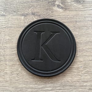 Puede incluir: Un posavasos redondo negro con un borde en relieve y una gran letra "K" en el centro. El posavasos est&aacute; hecho de un material mate y se encuentra sobre una superficie de madera.