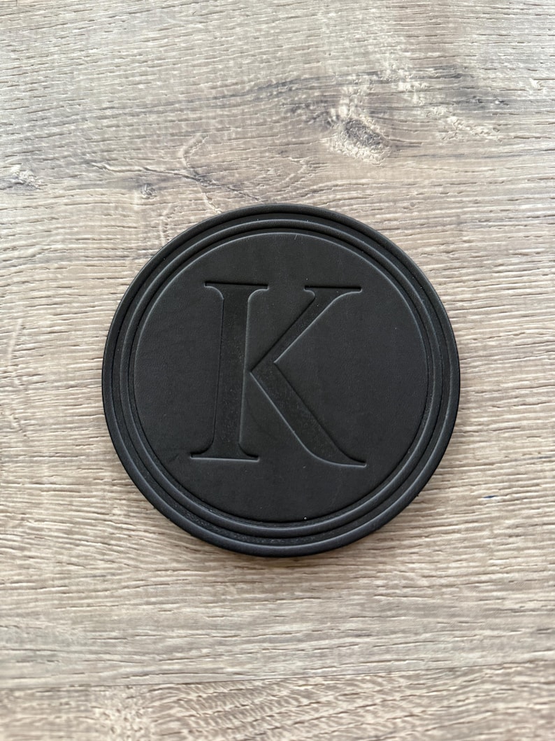 Puede incluir: Un posavasos redondo negro con un borde en relieve y una gran letra "K" en el centro. El posavasos est&aacute; hecho de un material mate y se encuentra sobre una superficie de madera.
