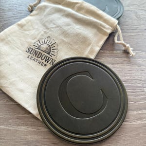 Puede incluir: Un juego de posavasos redondos de color gris oscuro con un borde en relieve y una gran letra "C" en el centro. Los posavasos est&aacute;n junto a una bolsa beige con cord&oacute;n con el logotipo "Sundown Leather". El fondo es una superficie de madera.