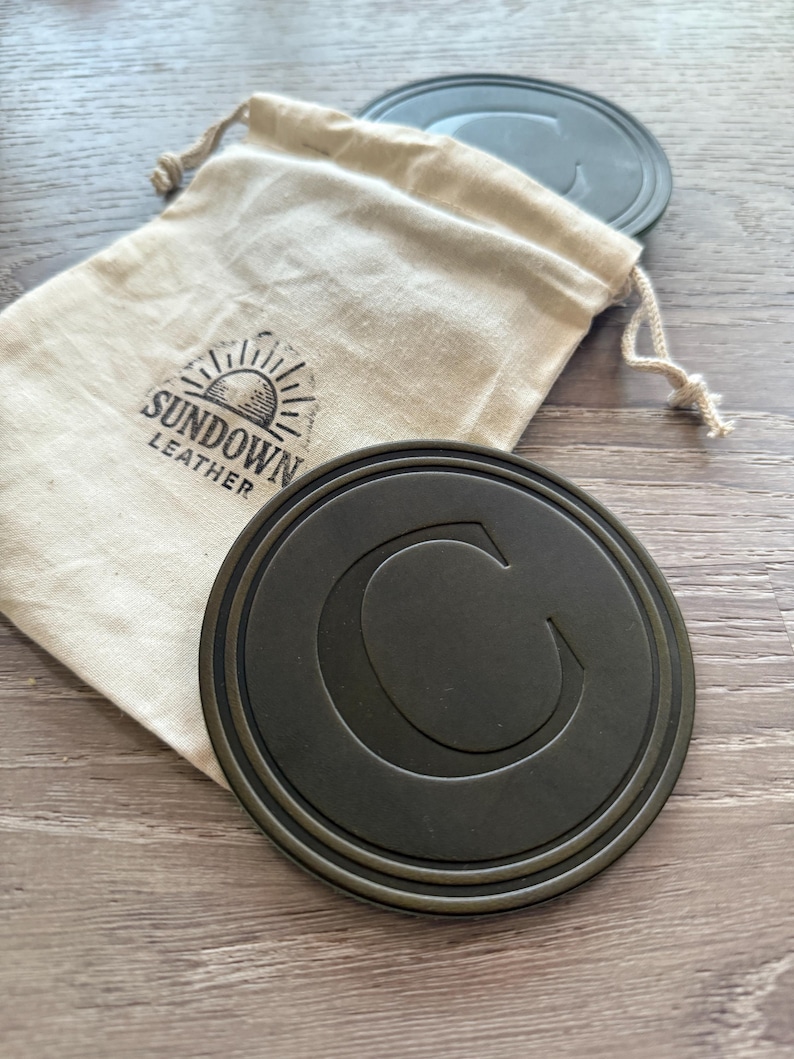 Puede incluir: Un juego de posavasos redondos de color gris oscuro con un borde en relieve y una gran letra "C" en el centro. Los posavasos est&aacute;n junto a una bolsa beige con cord&oacute;n con el logotipo "Sundown Leather". El fondo es una superficie de madera.