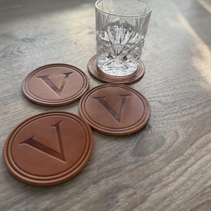 Puede incluir: Un vaso transparente con dibujos y cuatro posavasos de cuero marr&oacute;n con la letra "V" en relieve. Los posavasos son redondos y tienen un borde en relieve. El vaso est&aacute; sobre uno de los posavasos.