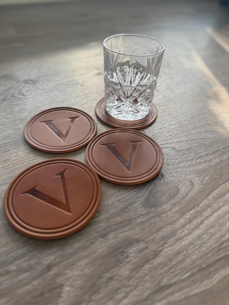 Puede incluir: Un vaso transparente con dibujos y cuatro posavasos de cuero marr&oacute;n con la letra "V" en relieve. Los posavasos son redondos y tienen un borde en relieve. El vaso est&aacute; sobre uno de los posavasos.