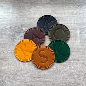 Puede incluir: Un conjunto de siete posavasos redondos de varios colores, cada uno con una letra del alfabeto. Los colores incluyen negro, marrón, verde oliva, naranja y verde oscuro. Las letras están en relieve.