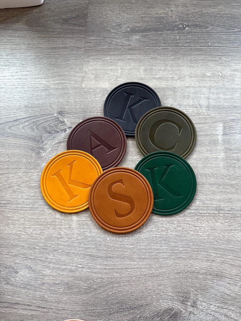 Puede incluir: Un conjunto de siete posavasos redondos de varios colores, cada uno con una letra del alfabeto. Los colores incluyen negro, marr&oacute;n, verde oliva, naranja y verde oscuro. Las letras est&aacute;n en relieve.