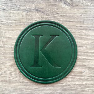 Puede incluir: Un posavasos redondo verde oscuro con un borde negro. La letra "K" est&aacute; grabada en el centro del posavasos. El posavasos est&aacute; dise&ntilde;ado para proteger las superficies de las marcas de bebidas.