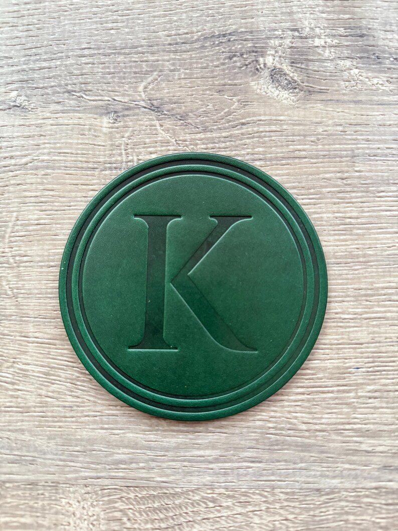 Puede incluir: Un posavasos redondo verde oscuro con un borde negro. La letra "K" est&aacute; grabada en el centro del posavasos. El posavasos est&aacute; dise&ntilde;ado para proteger las superficies de las marcas de bebidas.