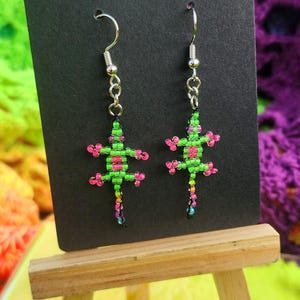 May include: A pair of beaded lizard earrings. The earrings are green with pink accents and dangle from silver-colored hooks. The earrings are displayed on a black card, propped up on a small wooden easel, against a colorful background.