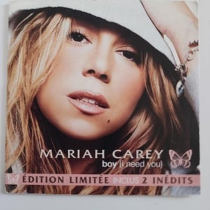 Puede incluir: Una portada de CD de música cuadrada con un retrato de Mariah Carey usando un sombrero. El texto dice MARIAH CAREY boy (i need you) y ÉDITION LIMITÉE INCLUS 2 INÉDITS con iconos de mariposas. La paleta de colores incluye marrón, blanco y rosa suave.