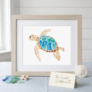 May include: A framed watercolor print of a sea turtle with a blue and teal shell and tan patterned skin. The light wood frame includes a white mat. A small card in front displays the text The Coastal Wildflower with a small flower illustration.