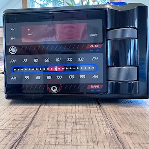 Puede incluir: Un radio despertador GE negro AM/FM con pantalla digital y dial de sintonización. La radio tiene un dial iluminado azul y rojo, controles de volumen y sintonización, y una toma de auriculares. El reloj está sobre una superficie de madera.
