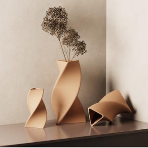 May include: Three beige ceramic vases with a twisted design. The largest vase holds dried flowers. The vases are arranged on a grey surface against a neutral background. The vases have a modern, minimalist aesthetic.