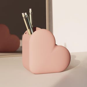 May include: A pale pink heart-shaped desk tidy containing several pencils and pens. The organiser is made of a smooth material and sits on a white surface. The pencils and pens are various colours.