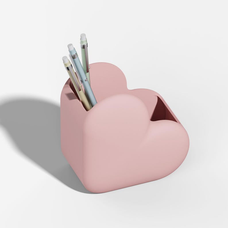 May include: A pink heart-shaped desk organizer holding three pens. The organizer is a solid, soft pink color and has a smooth surface. The pens are light blue, green, and yellow.