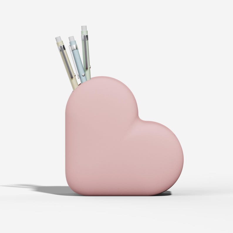 May include: A pink heart-shaped desk organizer holding three pens. The pens are light green, light blue, and silver. The organizer is made of a smooth material and sits on a white surface.