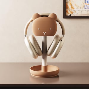 Könnte beinhalten: Ein brauner Bären-Kopfhörerständer mit weißem Stiel und Sockel, der ein Paar cremefarbene Kopfhörer hält. Der Ständer hat ein minimalistisches Design mit rundem Sockel und einem Bärengesicht an der Spitze.