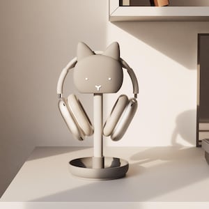 Pode incluir: Um suporte para fones de ouvido cinza claro em forma de coelho, com um par de fones de ouvido cinza claro. O suporte tem uma base redonda e um poste vertical que suporta a cabeça do coelho. Os fones de ouvido estão apoiados no suporte, com os fones voltados para a frente.