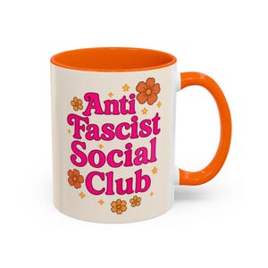 Może przedstawiać: Biały ceramiczny kubek z pomarańczowym wnętrzem i uchwytem. Kubek zawiera napis "Anti Fascist Social Club" w kolorze różowym, z pomarańczowymi akcentami kwiatowymi. Do gorących napojów.