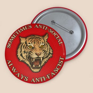 May include: A red button featuring a tiger's head and the text "SOMETIMES ANTI-SOCIAL ALWAYS ANTI-FASCIST". The tiger is depicted with an open mouth. The button has a silver pin on the back.