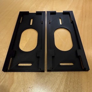 Puede incluir: Dos componentes de plástico negros y rectangulares con un recorte ovalado y varias ranuras rectangulares. Los componentes tienen forma trapezoidal y están diseñados para entrelazarse. Los artículos están colocados sobre una superficie de madera de color marrón claro.