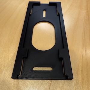 Puede incluir: Soporte de plástico negro rectangular con un recorte ovalado en el centro. El soporte tiene forma trapezoidal con ranuras y pestañas en los bordes. La superficie tiene un patrón texturizado y acanalado.