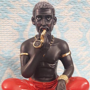 May include: A black figurine of a seated figure with red pants, gold jewelry, and a gold pipe. The figure is seated on a gold base. The figurine has the text "Papacandelo" on the base.