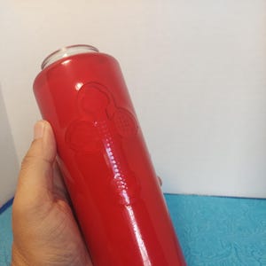 Può includere: Un barattolo di candela in vetro rosso con un disegno a croce. Il barattolo è cilindrico con un coperchio in vetro trasparente. La croce è in rilievo sulla parte anteriore del barattolo. Il barattolo di candela è tenuto da una persona.