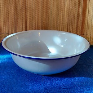 Puede incluir: Un cuenco de esmalte blanco con borde azul. El cuenco, de forma ovalada, se apoya sobre una superficie azul estampada. El fondo presenta una estera tejida de color beige.