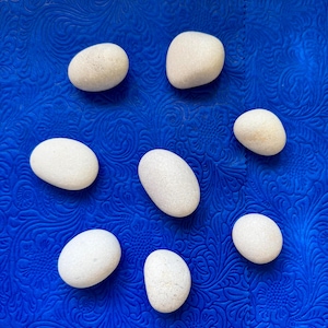 May include: Seven smooth, rounded, off-white stones are arranged in a circular pattern. Each stone has a matte texture and a solid, light cream color. The stones vary slightly in size and shape, appearing as natural, polished river rocks.