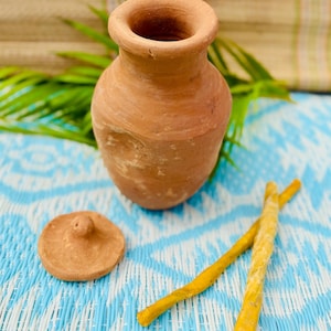 May include: A small, unglazed terracotta vase with a wide opening and a matching lid. Two yellow, twisted sticks are next to the vase on a blue and white patterned surface. The vase is a warm, earthy brown color.
