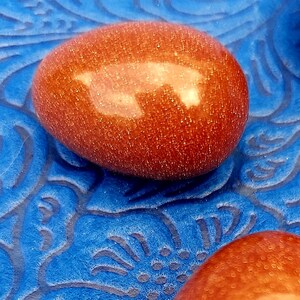 May include: A close-up of a polished, oval-shaped, orange-red gemstone with a reflective surface. The gemstone sits on a textured, deep blue surface with a swirling pattern. The stone has a unique, natural appearance.