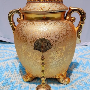 Könnte beinhalten: Eine goldfarbene dekorative Urne mit floralem Muster, verzierten Griffen und einem kronenförmigen Deckel. Vor der Urne befindet sich eine kleine goldene Glocke mit dekorativem Griff. Die Urne steht auf einer blau-weiß gemusterten Oberfläche.