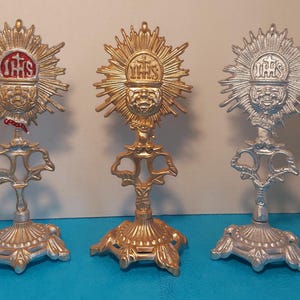 Könnte beinhalten: Drei verzierte Monstranzen in Gold- und Silbertönen. Jede hat ein Sonnenstrahlendesign mit den Buchstaben IHS, einen dekorativen Sockel und einen detaillierten Ständer. Die Monstranzen werden auf einer blauen Oberfläche präsentiert.