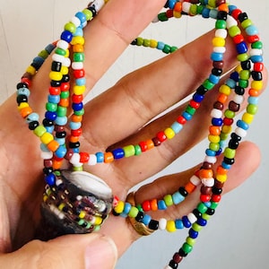 May include: A colorful beaded necklace with a large, decorative pendant. The necklace features strands of small, round beads in various colors, including blue, green, yellow, red, and white. The pendant is a dark, textured shape with colorful accents.