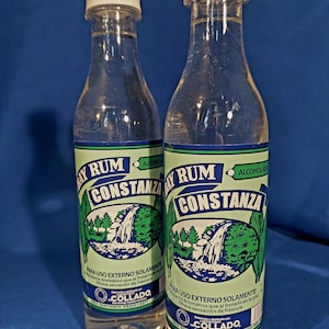 May include: Two clear bottles of Bay Rum Constanza, each with a white cap and a green and blue label. The label features a waterfall scene. The bottles are filled with a clear liquid.